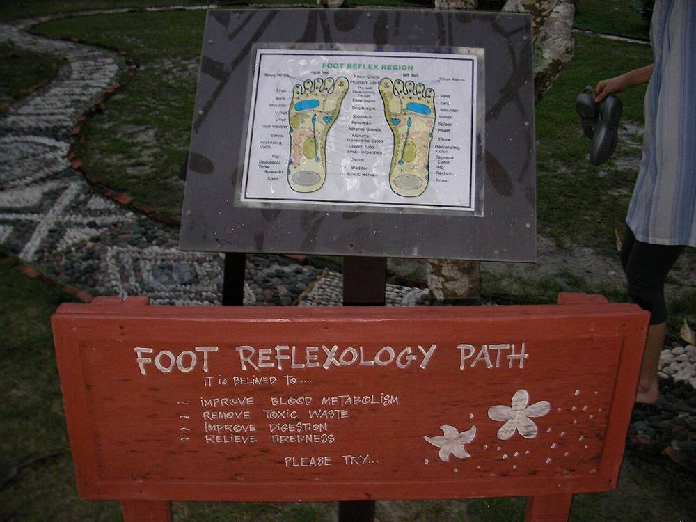 A foot reflexology path sign with a foot reflex chart beside a pebble walkway at a Malaysian resort