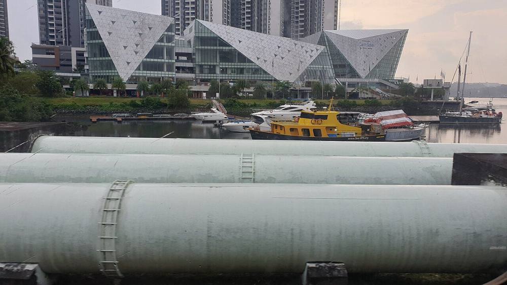 Large water pipes running along the Causeway between Johor Bahru and Singapore, with a marina and glass buildings behind