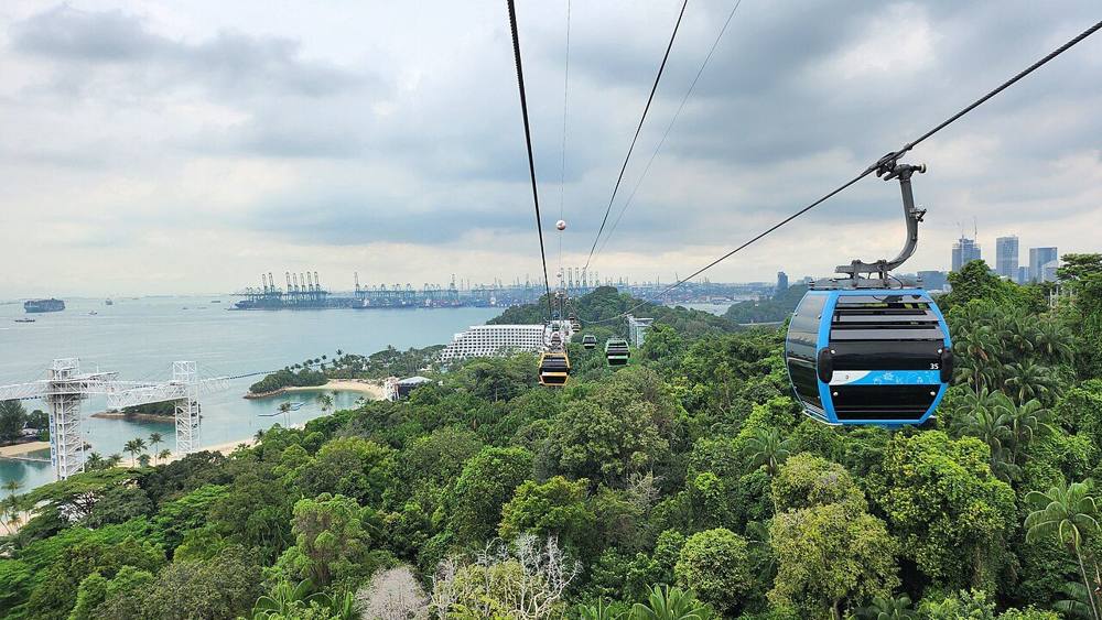 A cable car gondola over the green forest of Sentosa with the sea and port cranes in the distance
